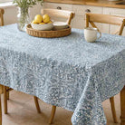 A rectangular table covered with a light blue and white patterned tablecloth, set in a bright kitchen with wooden chairs, a wicker tray with lemons, stacked plates, and a ceramic mug.