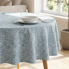 A round table covered with a light blue and white patterned tablecloth, set with a white bowl, plate, glass, and silverware in a bright room near a window.