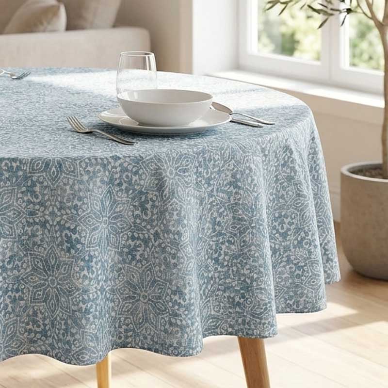 A round table covered with a light blue and white patterned tablecloth, set with a white bowl, plate, glass, and silverware in a bright room near a window.