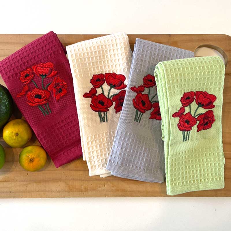 Four embroidered kitchen towels with red floral poppy designs on a wooden surface.