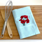 White towel with red floral embroidery on a wooden cutting board with a whisk and spatula.
