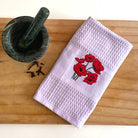 White towel with red floral embroidery on a wooden surface next to a green mortar and pestle.