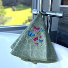 A sage green textured hand towel with embroidered colorful butterflies, draped over a chrome faucet on a white bathtub ledge with a window and outdoor view in the background.