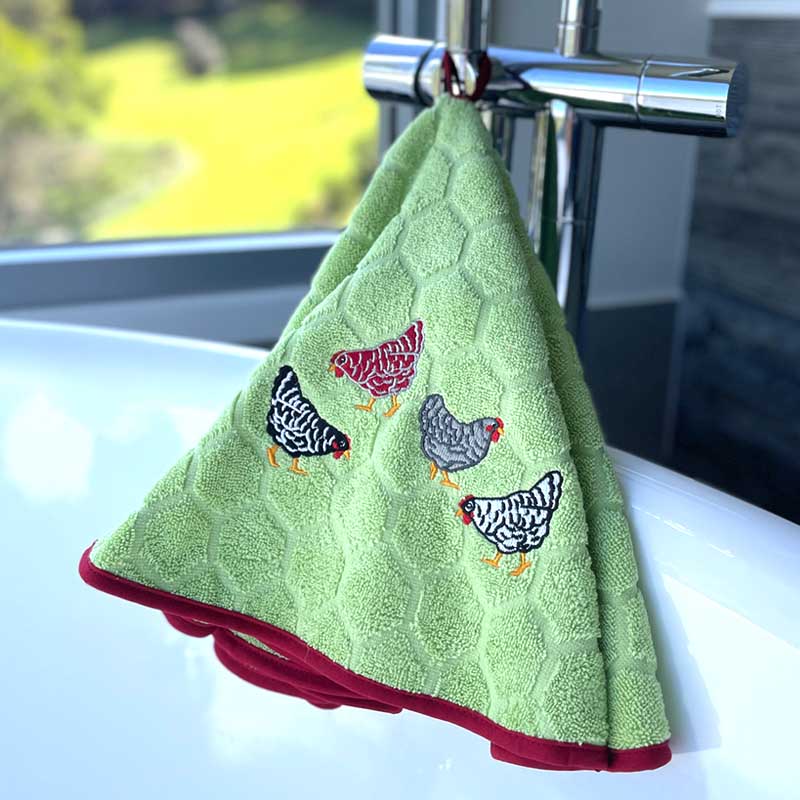 A light green textured kitchen towel with embroidered chickens and a dark red border, hanging from a chrome faucet over a white sink.
