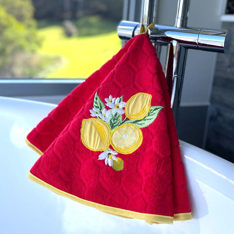 A red textured kitchen towel with embroidered lemons, flowers, and green leaves, hanging from a chrome faucet in a bright kitchen setting.