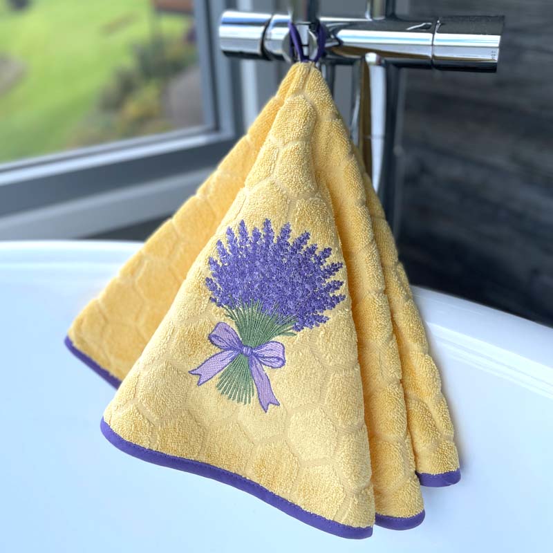 Round hand towel in yellow with lavender embroidery and purple trim hanging on a loop. Positioned hanging on the tap of a spa bath.