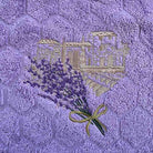 A close-up of a lavender-themed hand towel featuring a textured purple background with a raised floral pattern, embroidered lavender sprigs, and a gold-toned bastide motif.