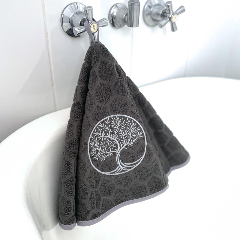 A grey hand towel with a silver embroidered tree of life design, hanging from a chrome wall hook above a white bathtub.