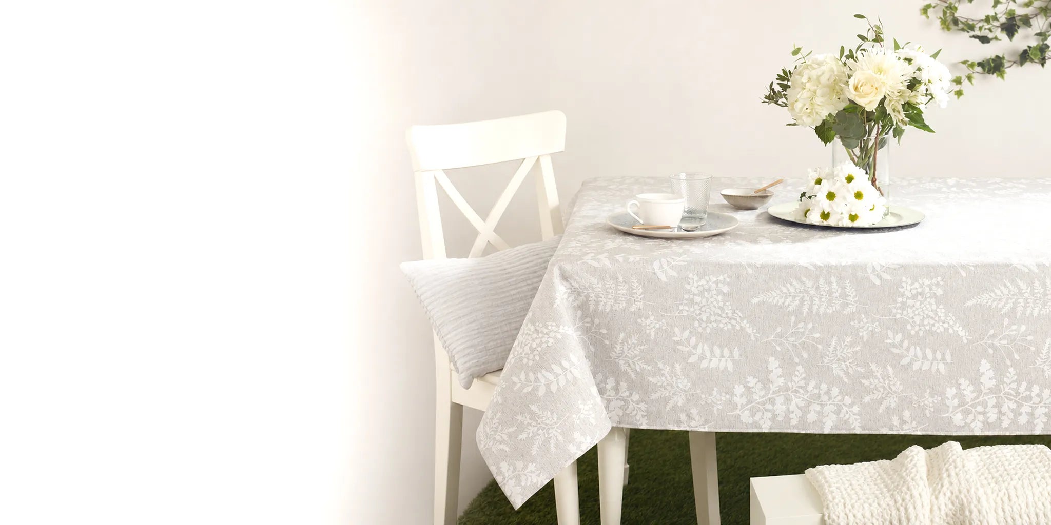 A stain-resistant tablecloth with a delicate gray and white floral pattern draped over a dining table, styled with a white chair, a vase of white flowers, and a teacup on a saucer.