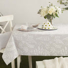 A light gray tablecloth with a white botanical leaf pattern draped over a dining table, styled with a white floral vase, a teacup and saucer, and a small bowl.