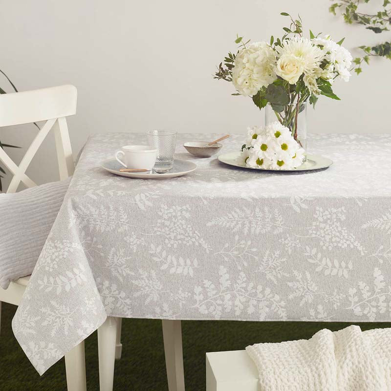 A light gray tablecloth with a white botanical leaf pattern draped over a dining table, styled with a white floral vase, a teacup and saucer, and a small bowl.